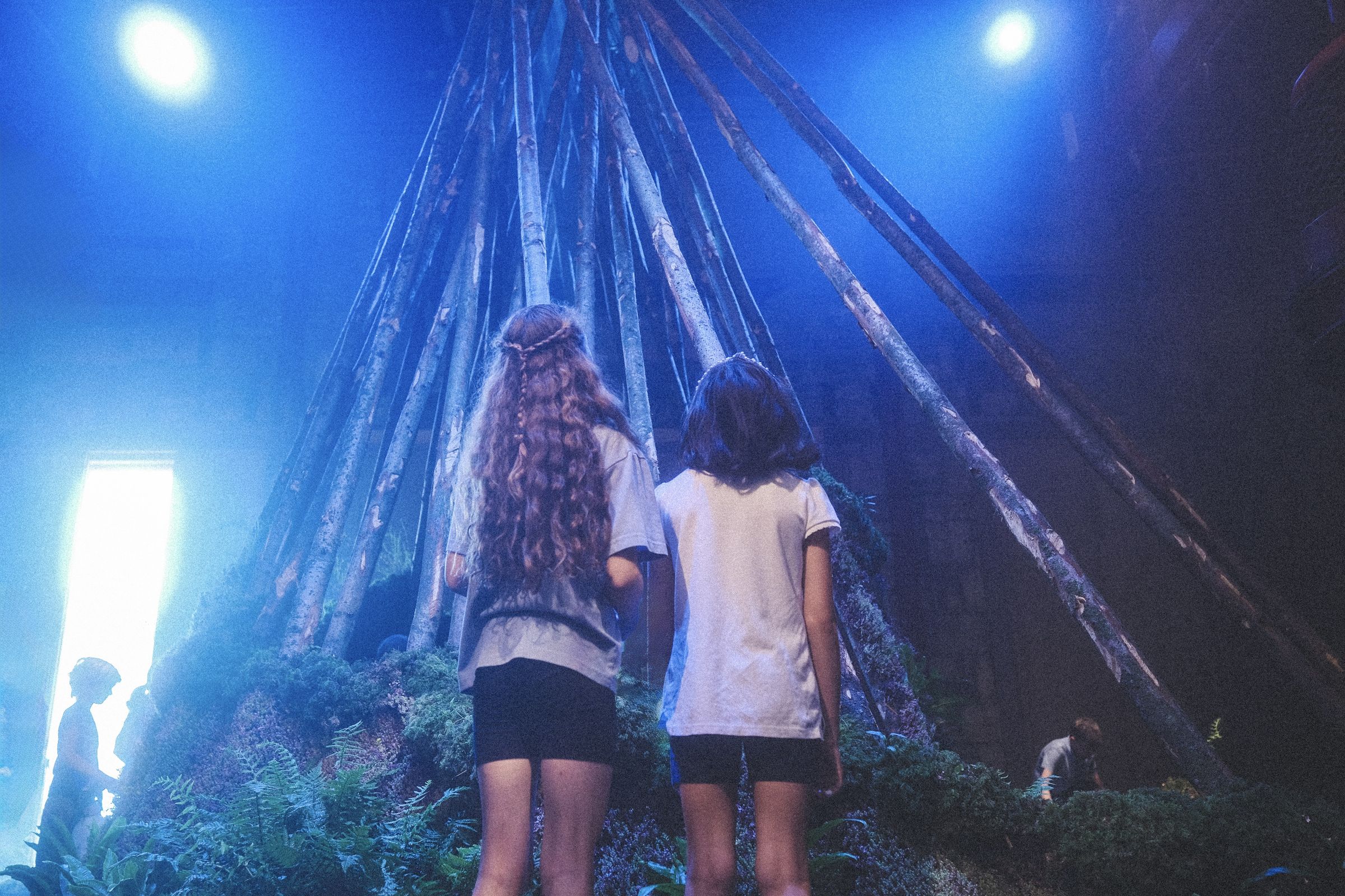 Two girls, seen from behind, look up into the Living Tree, a tepee of bare poles lit in deep blue, with bright light at the left.