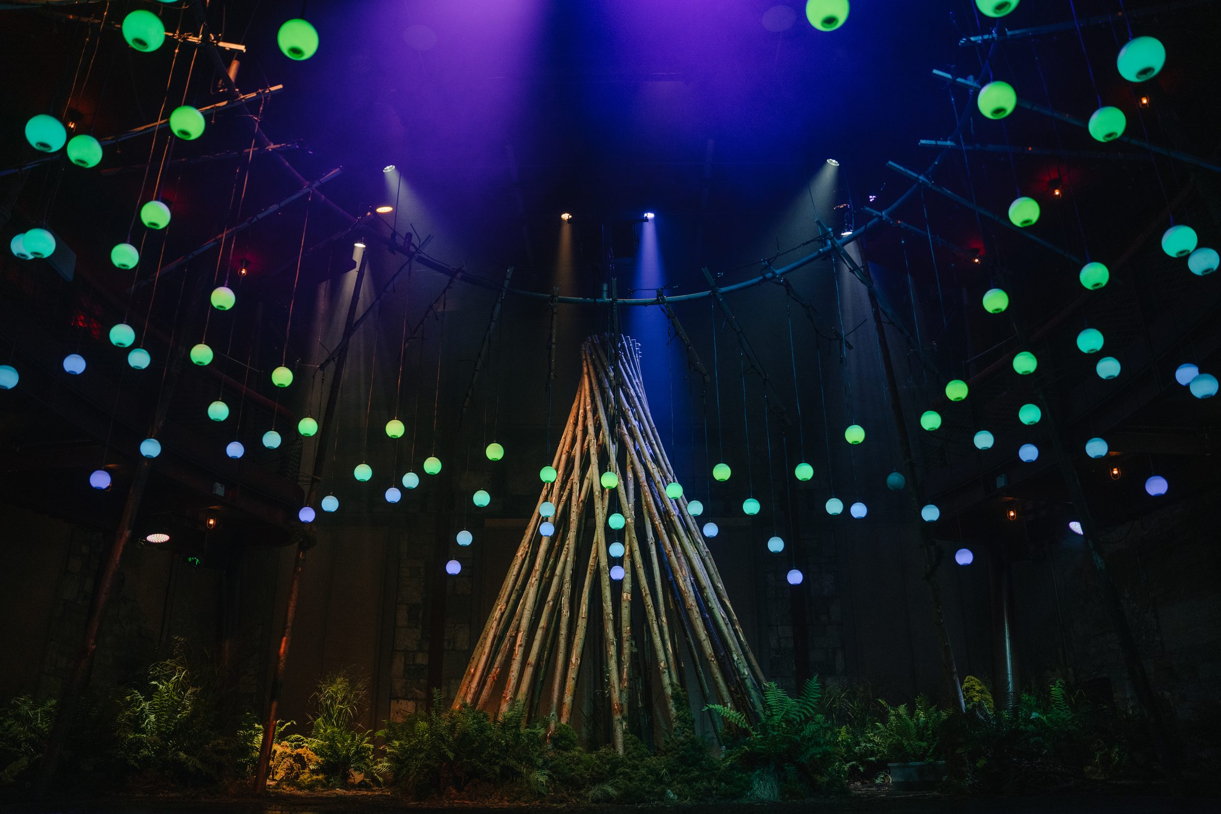 The Living Tree at The Egg, Theatre Royal Bath: a tall tepee of bare poles ringed with ferns, with glowing green and blue orbs hanging in the dark space around it