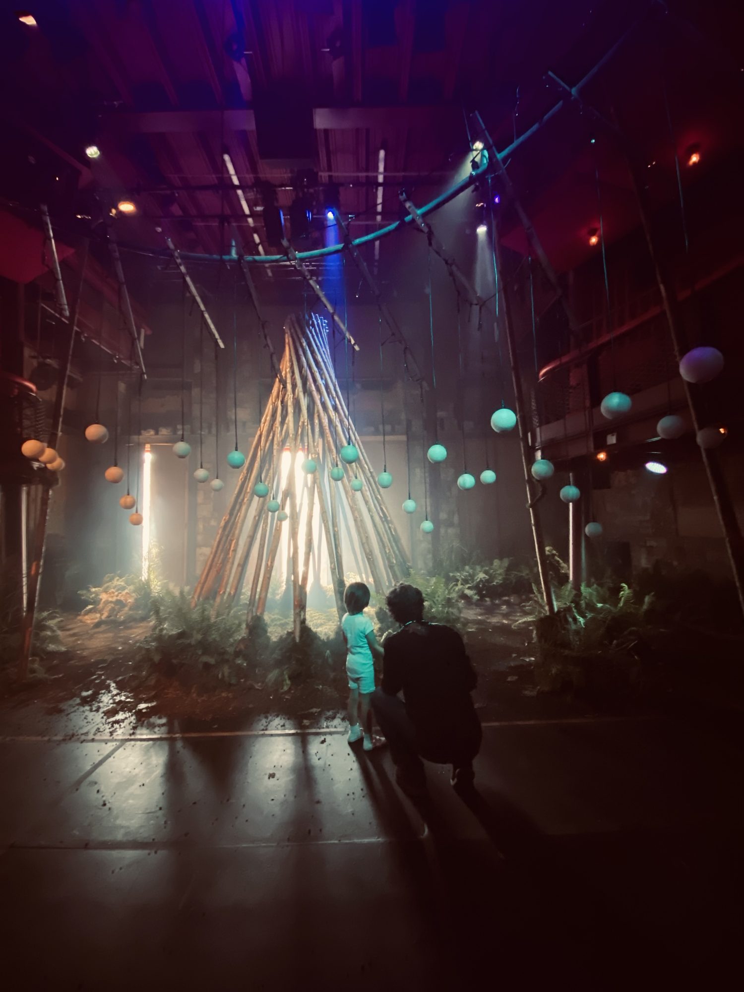 A child and an adult crouching beside them, seen from behind, look toward the Living Tree: a tepee of bare poles in a dark hall, ringed with ferns, with pale spheres hanging around it and bright light behind.