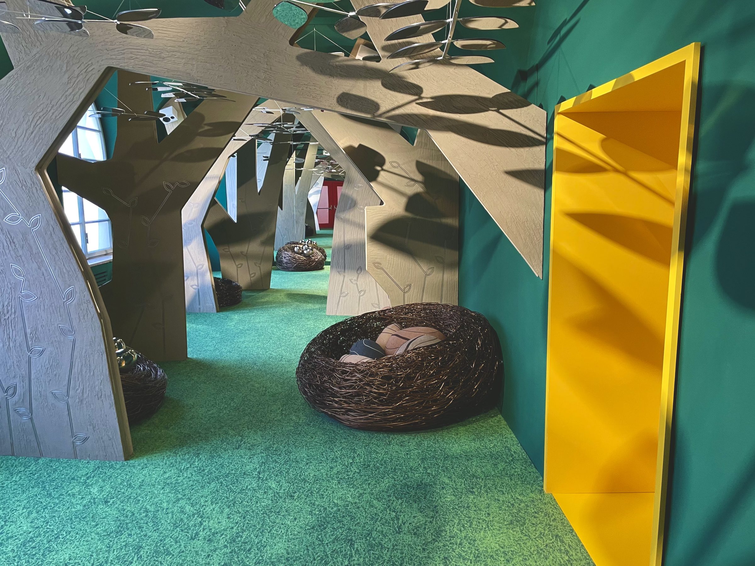 A corridor of tree-shaped wooden arches hung with mirrored leaf mobiles, woven nests on a green carpet and a yellow doorway