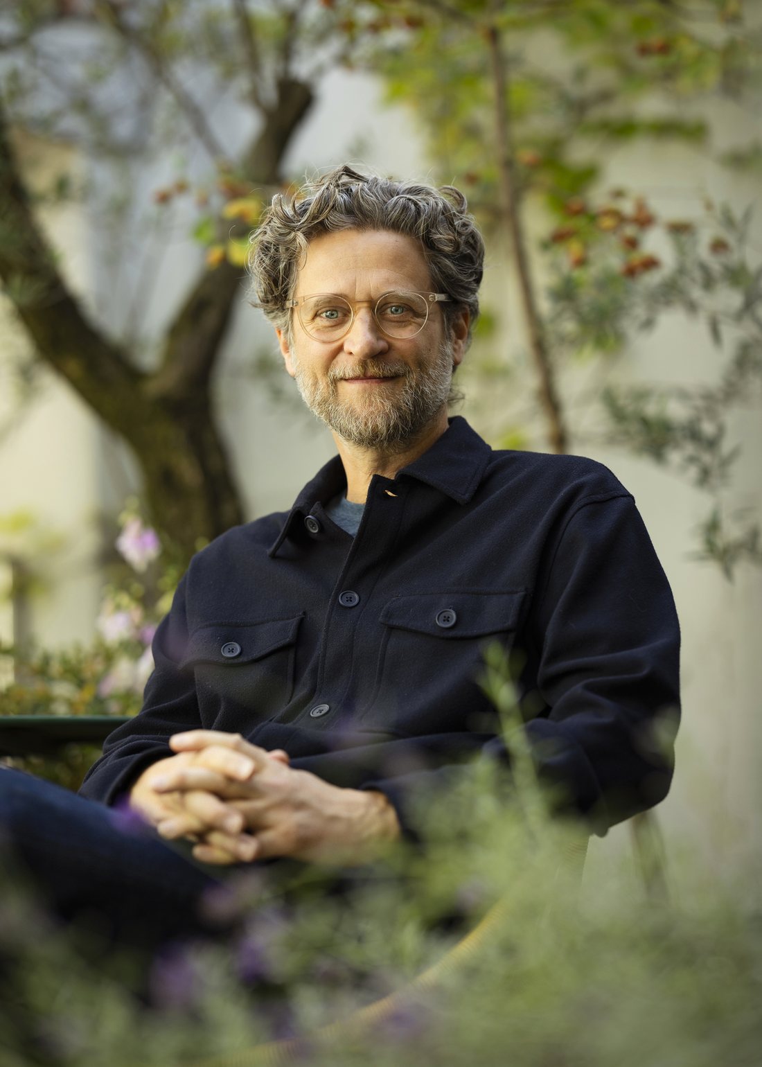 Portrait of Andrew Amondson, seated outdoors in a garden with his hands clasped, wearing round glasses and a dark jacket.